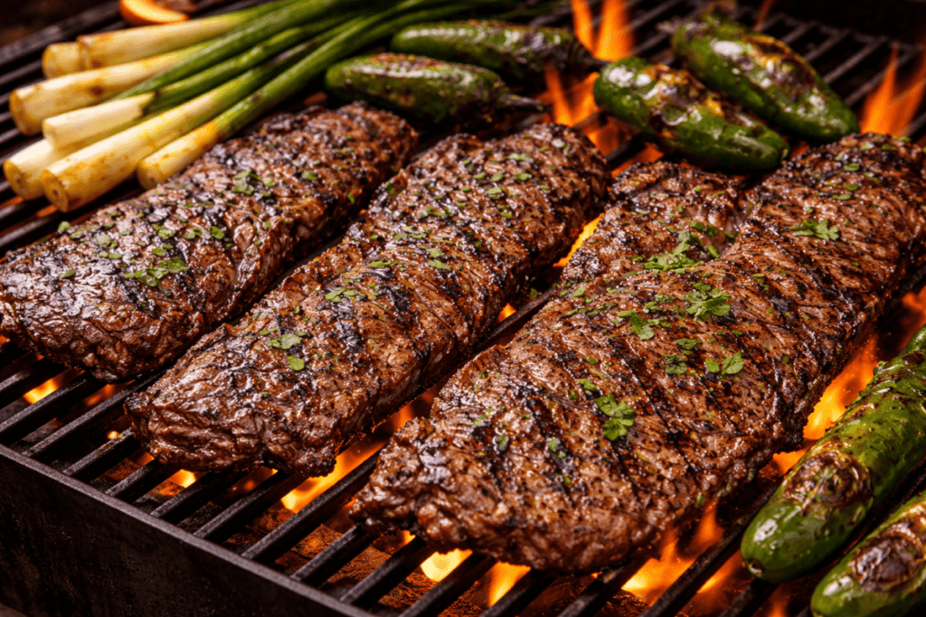 Carne asada cooking on a hot grill with marinated skirt steak sizzling over open flames alongside charred jalapeños and green onions.
