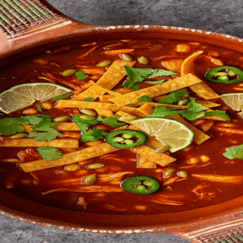 Sopa Azteca tortilla soup in a Mexican clay bowl with crispy tortilla strips, lime, cilantro, jalapeños, and pepitas