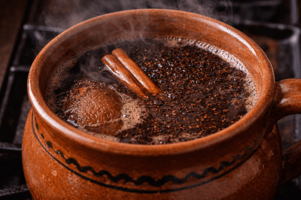 Clay pot of café de olla simmering on the stovetop with cinnamon sticks, piloncillo melting into dark coffee, and steam rising from the spiced brew.