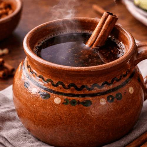 Traditional café de olla served in a rustic clay mug, filled with dark Mexican coffee and topped with a cinnamon stick, steam rising on a wooden table.