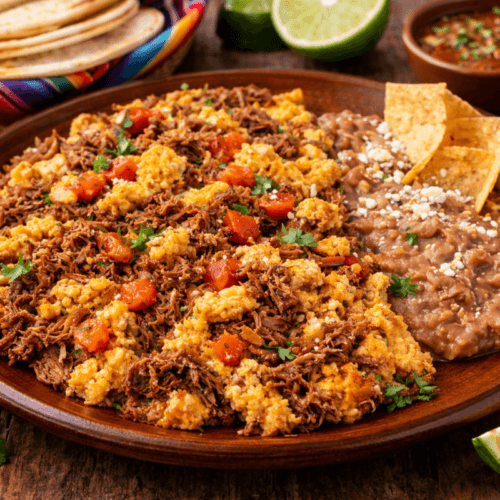 Machaca con huevo served on a rustic wooden table with flour tortillas, refried beans, and salsa