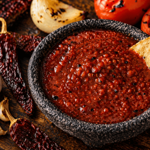 Chipotle salsa in a stone molcajete with dried chipotles, charred tomatoes, onion, and garlic on a rustic wooden surface.