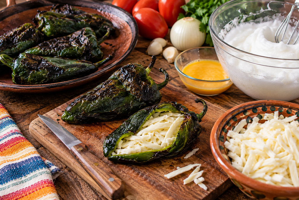 Roasted poblano peppers being stuffed with shredded Oaxaca cheese for traditional Mexican chiles rellenos, surrounded by whipped egg whites, egg yolks, and fresh ingredients on a rustic wooden cutting board.